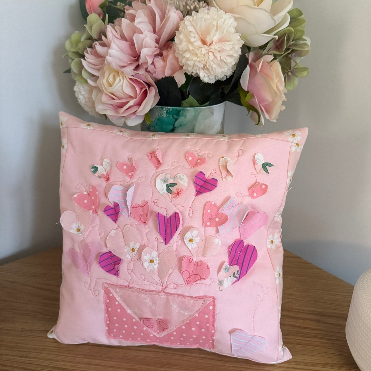 Pink decorative pillow with heart patterns on a wooden surface, with flowers in the background.