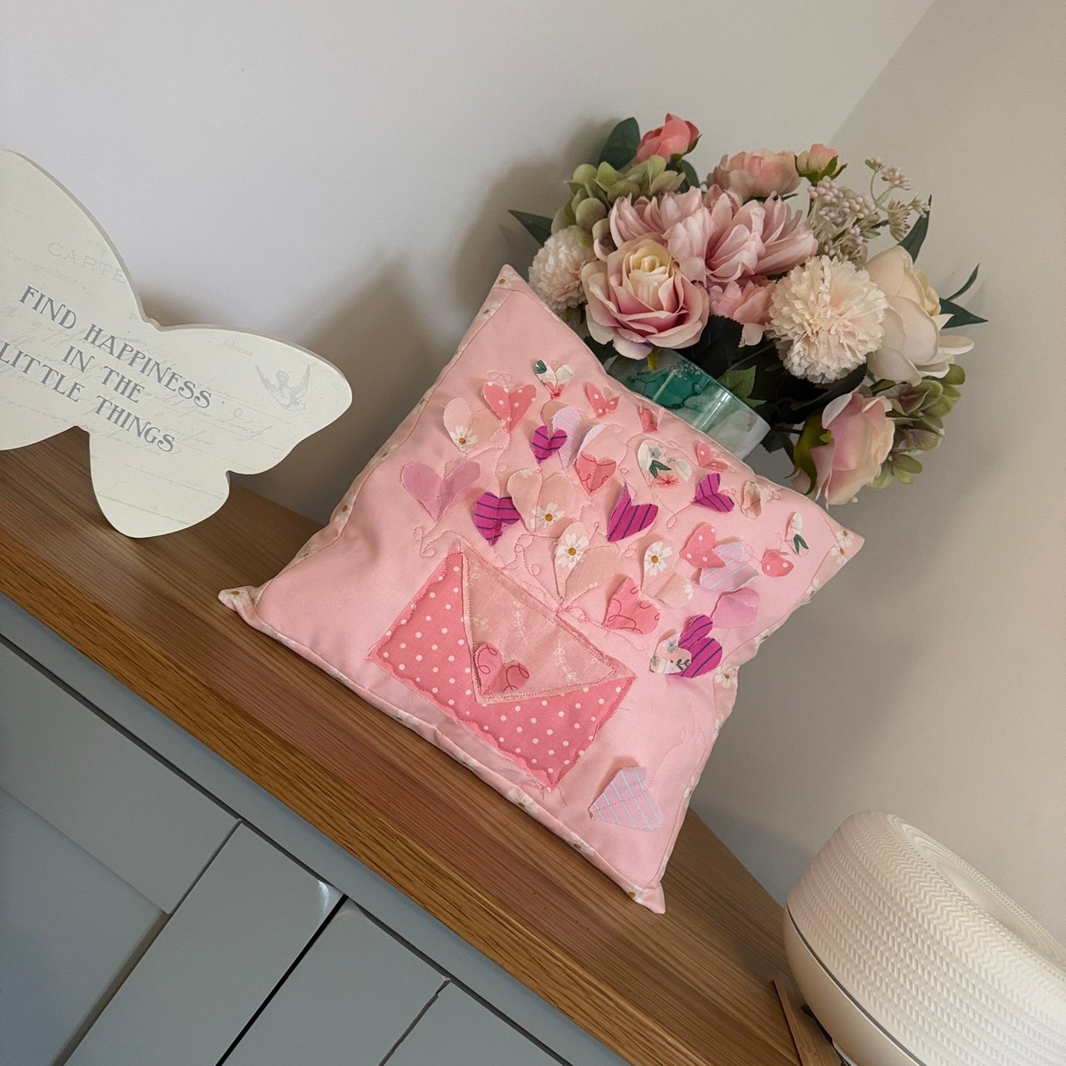 Pink decorative pillow with floral patterns on a wooden shelf with flowers and a decorative plate.