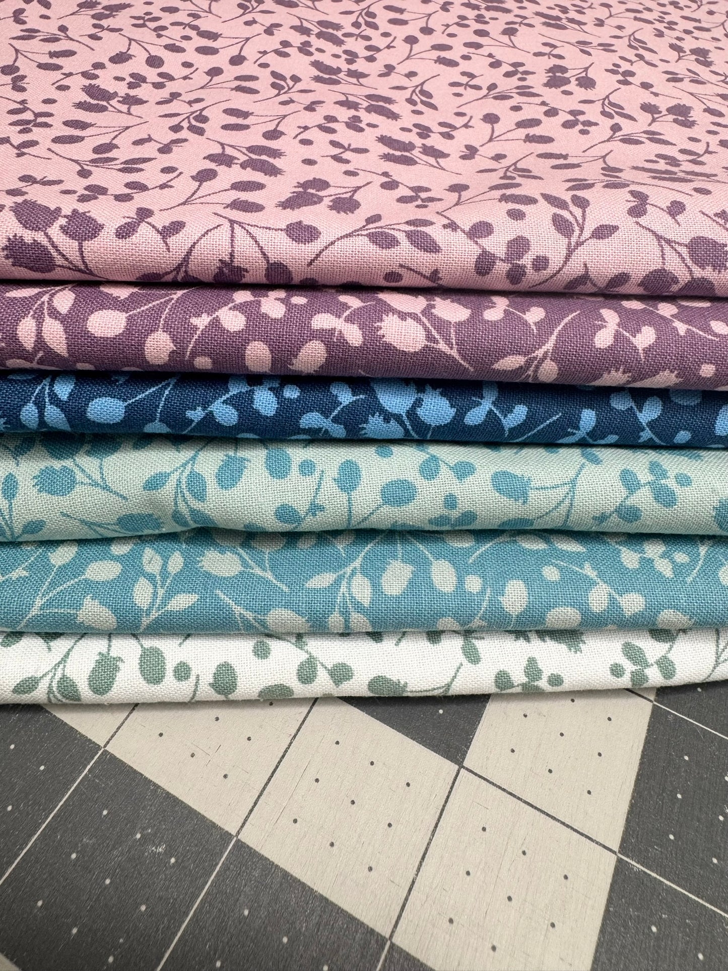Stack of fabric fat quarters for quilting with floral patterns in purple, blue, and green on a cutting mat.