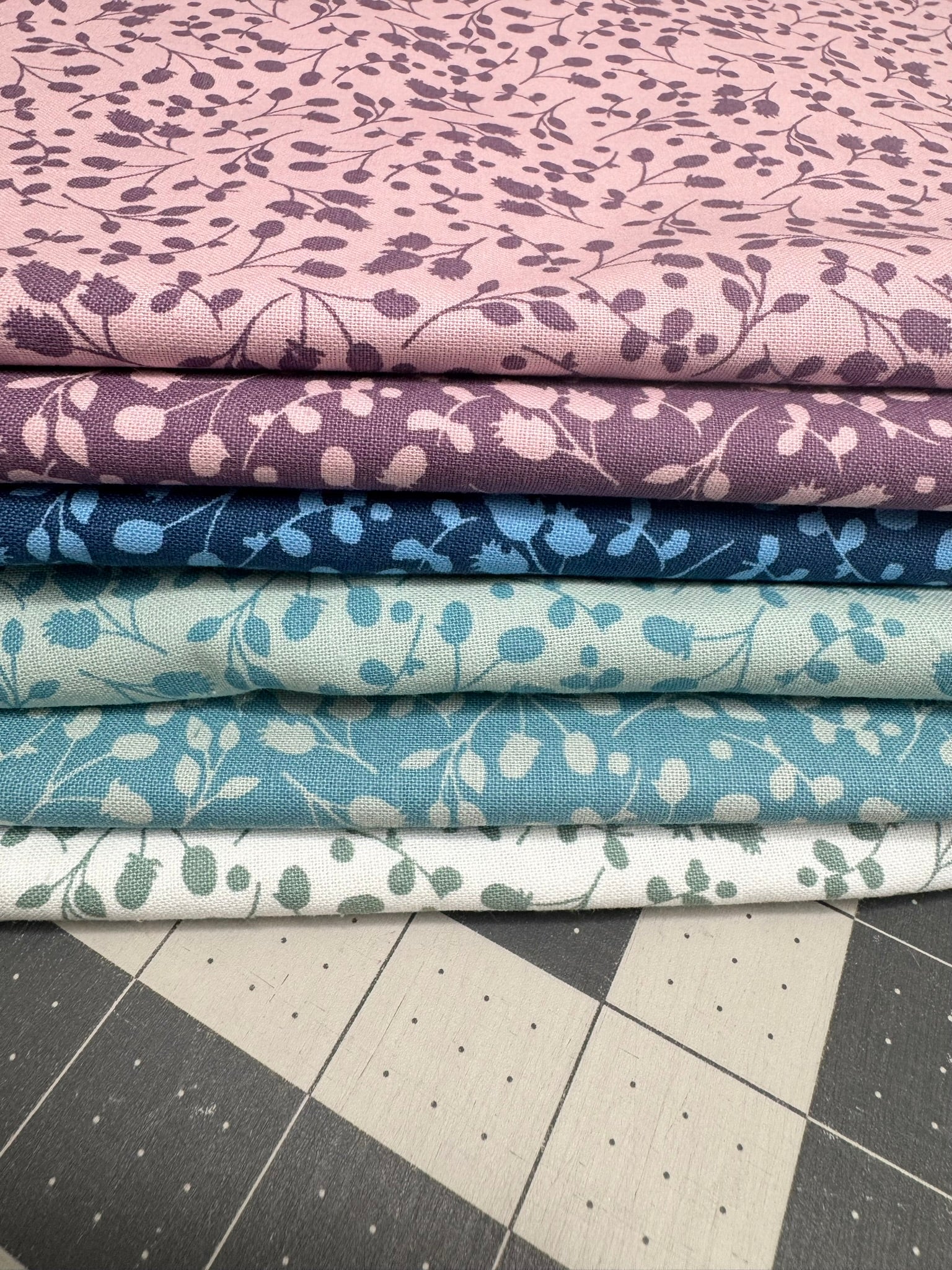 Stack of fabric fat quarters for quilting with floral patterns in purple, blue, and green on a cutting mat.